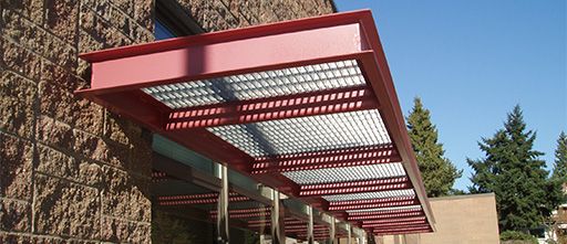 Photo of a red sun shade or canopy, placed on the side of a building, and has bar grating infill panels.